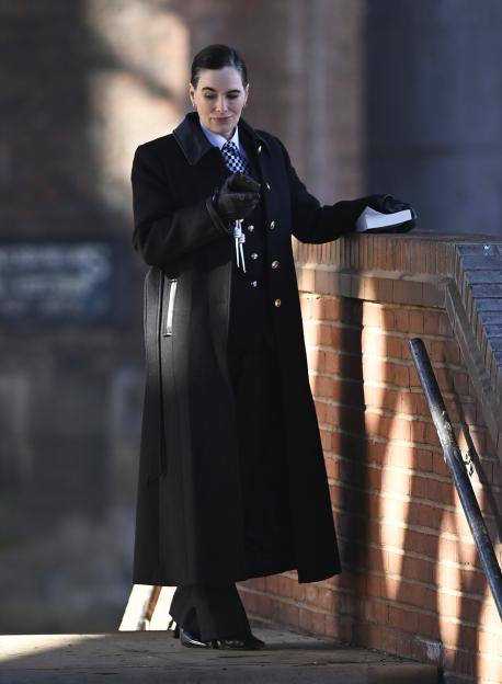 Lena Headey in character for "Project Codename" wearing a long black coat, a black and white checkered tie, and holding keys, while leaning on a brick wall.