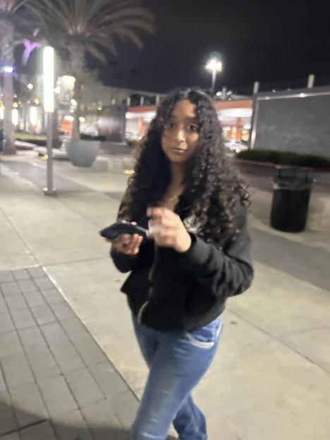 Celeste Rivas wearing a black hoodie and blue jeans, holding a phone, on a street at night.