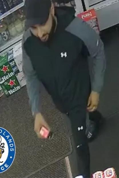 Mohammed Maneer Khan, wanted for murder, wearing a black and gray jacket, black pants, and a black baseball cap, holding a small object.