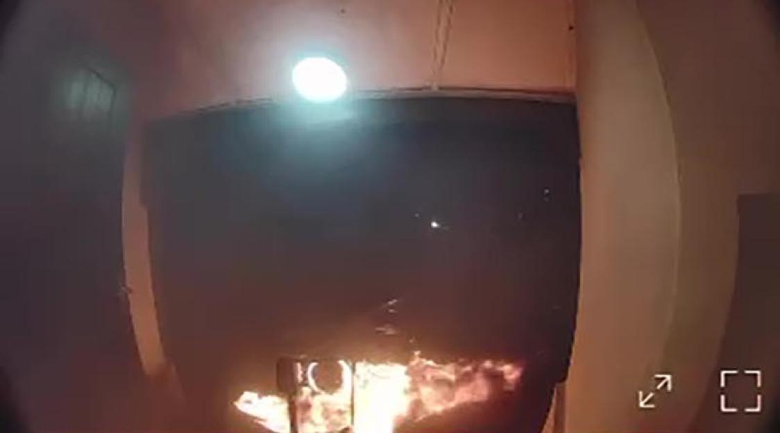 Security camera footage of an arson attack with flames visible at the bottom of the frame and smoke filling the air.