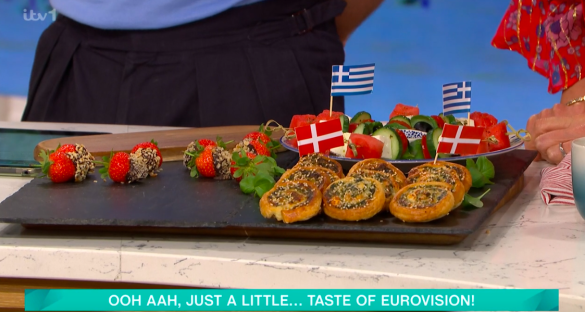 Screenshot of food representing Eurovision: chocolate-covered strawberries, savory pastries, and a Greek salad with Danish and Greek flags.