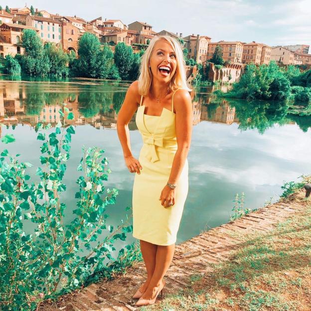 Laura Hamilton in a yellow dress by a river with a town in the background.