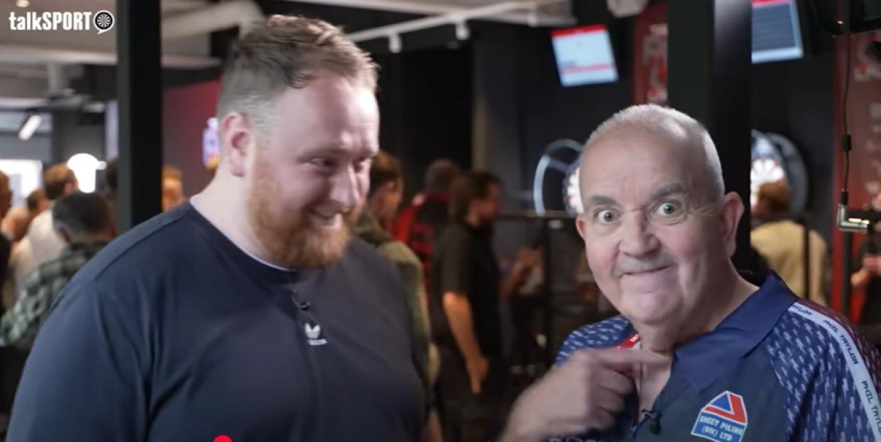 Two men in a darts venue, one smiling and one pointing to his shirt.