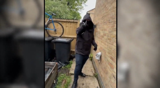 A hooded figure making a hand gesture in an alleyway.
