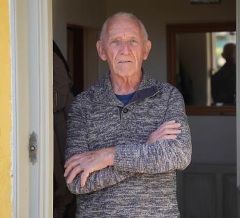 Older man standing in doorway with arms crossed.