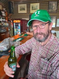 A man in a green Heineken cap sits at a bar holding a Heineken beer.