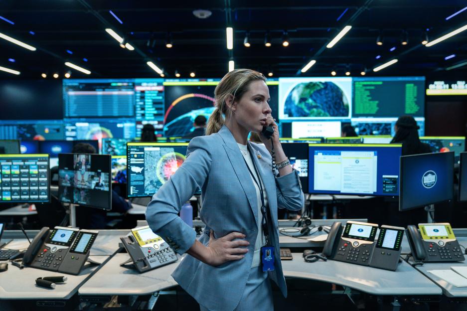 Olivia Walker in a light blue pantsuit talking on a black corded phone in a command center.