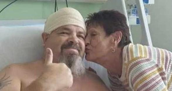 Man in hospital bed giving thumbs up, kissed on the cheek by a woman.