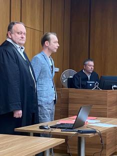 Three men in a courtroom. Two defendants stand before a judge.