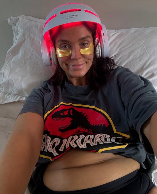Selfie of a woman wearing a red light therapy helmet and gold under-eye patches.