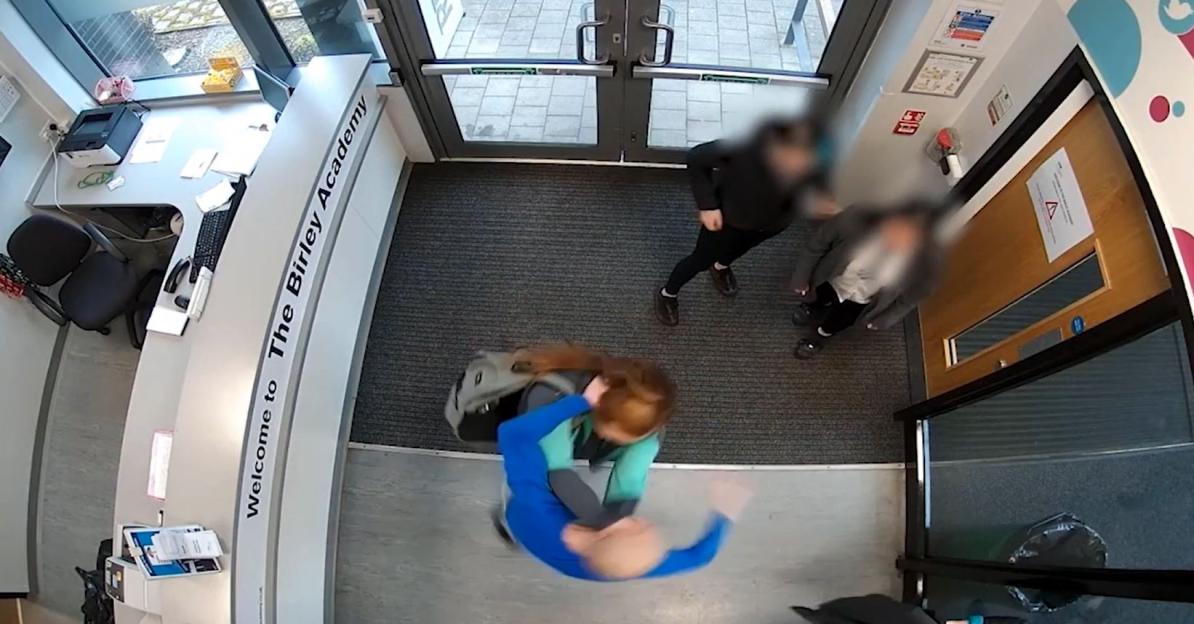 Overhead view of a student in a blue top and another in a mint green top facing a desk that reads "Welcome to The Birley Academy," while two other people stand in the background.