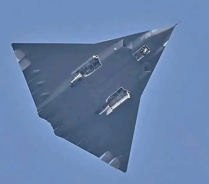 A dark grey stealth bomber flying in a clear blue sky, with its landing gear extended.