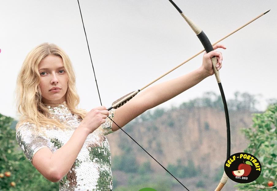 Apple Martin in a sequin dress holding a bow and arrow in an apple orchard, with "Self-Portrait" brand stickers.