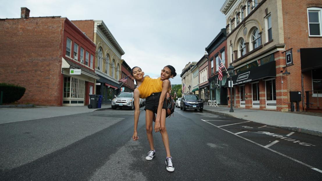 Conjoined twin sisters Carmen and Lupita smiling at the camera in the middle of a street lined with buildings.