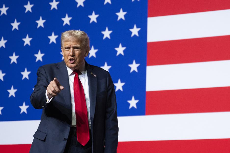 Donald Trump pointing at the viewer in front of an American flag.