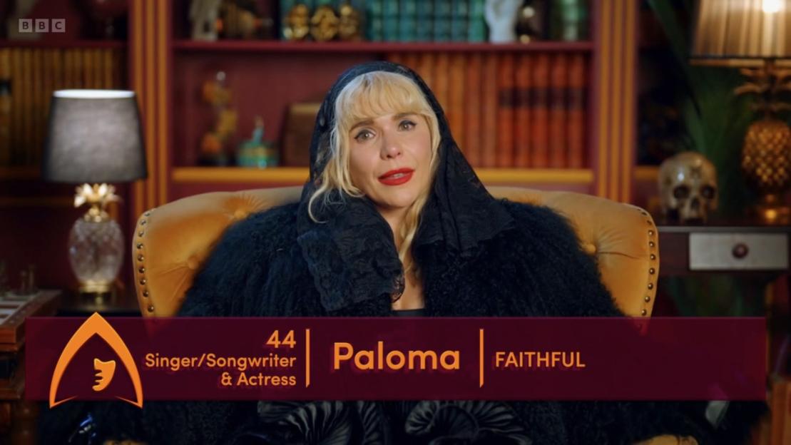 Paloma Faith sitting on a mustard yellow chair in a black lace veil and black fluffy top.