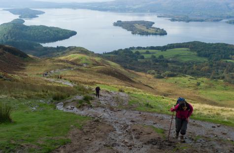 Hikers descending a muddy trail overlooking Loch Lomond in Scotland.