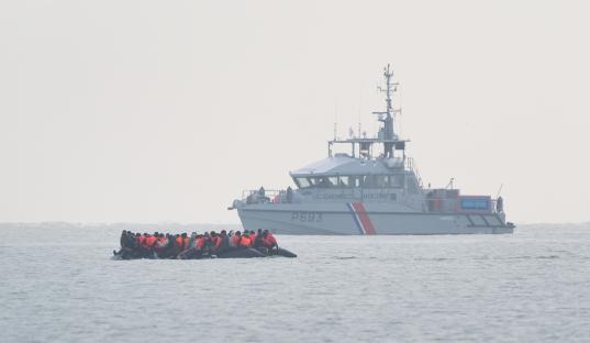 Migrants in a small boat being monitored by a larger vessel.
