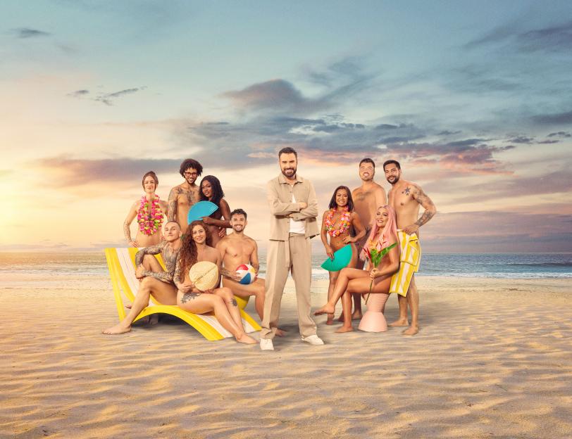 Group photo of a reality show's cast and host on a beach.