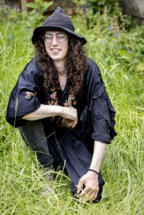 Person with long curly hair, glasses, and a black hat sitting in the grass.