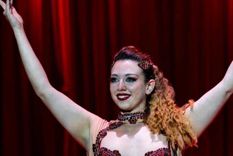 A woman in a red, jeweled costume with arms raised and curly hair smiling on a stage.