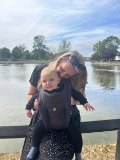 A mother holding her baby in a carrier by a pond.