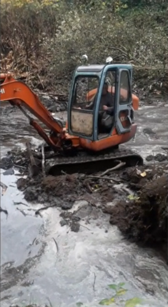 A small orange excavator with a grey cabin in a muddy, shallow stream.
