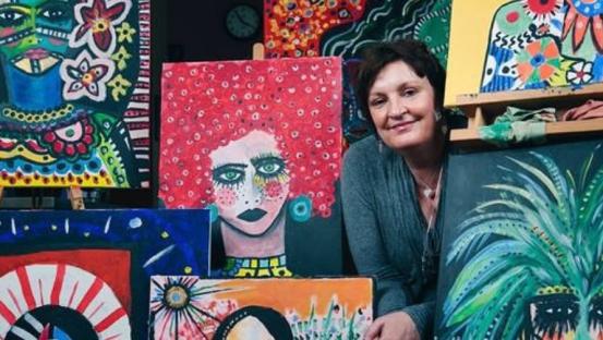 A woman sits surrounded by her vibrant, colorful paintings.