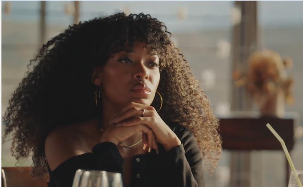 A woman with curly hair looks thoughtfully into the distance, her hands clasped under her chin.