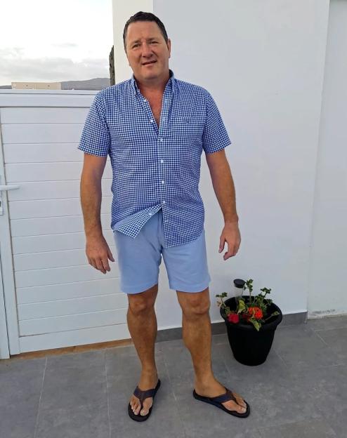 A man in a blue and white gingham shirt and light blue shorts stands outside.