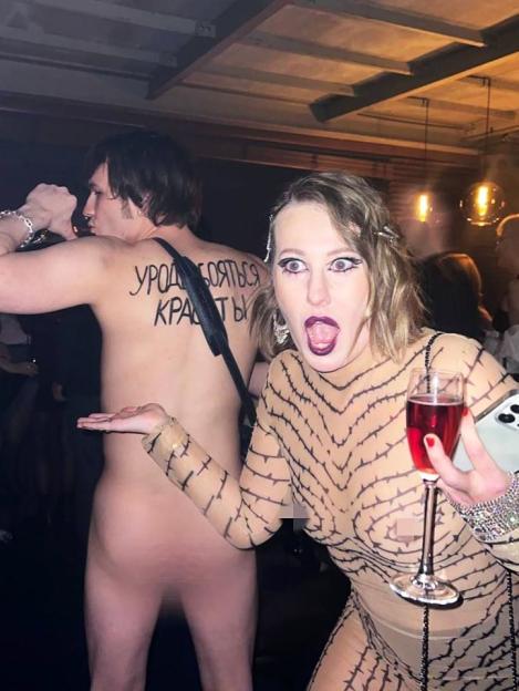 Woman in a sheer, thorn-patterned bodysuit holding a glass of champagne, with a naked man in the background; text is visible on the man's back.