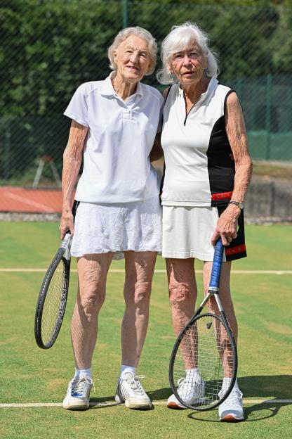 Two elderly women, tennis partners, stand on a tennis court holding rackets.