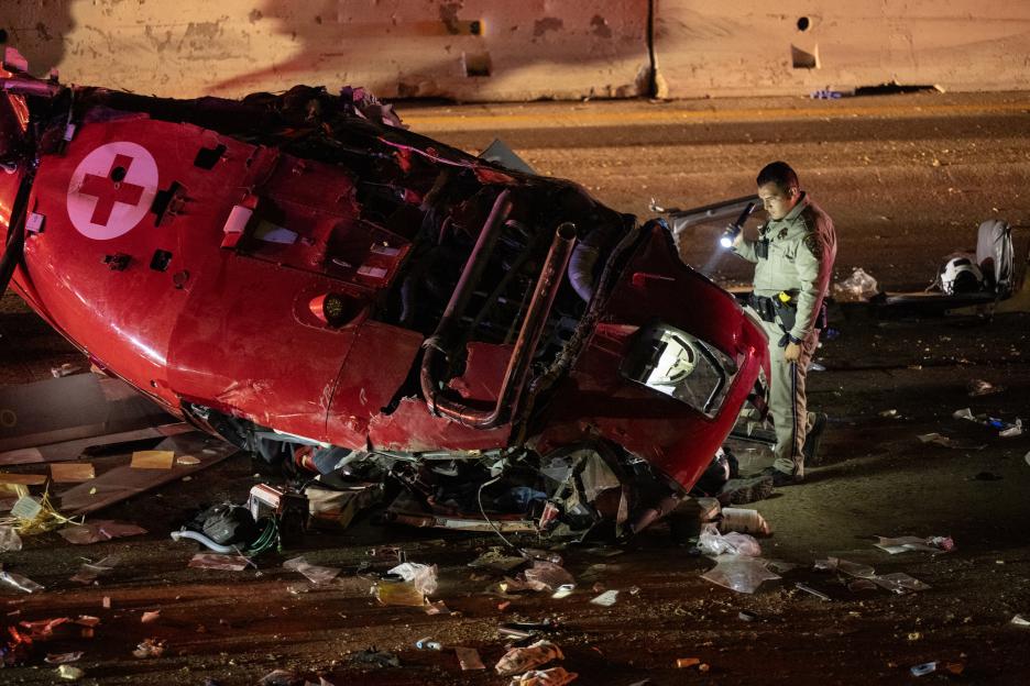 A red medevac helicopter is split down the middle on a highway with debris scattered around, as a CHP investigator uses a flashlight to search.