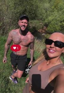 Couple posing outdoors; man shirtless, woman in athletic wear.