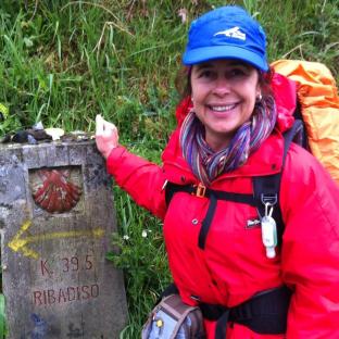 Woman in red jacket and blue hat by a Ribadiso trail marker.