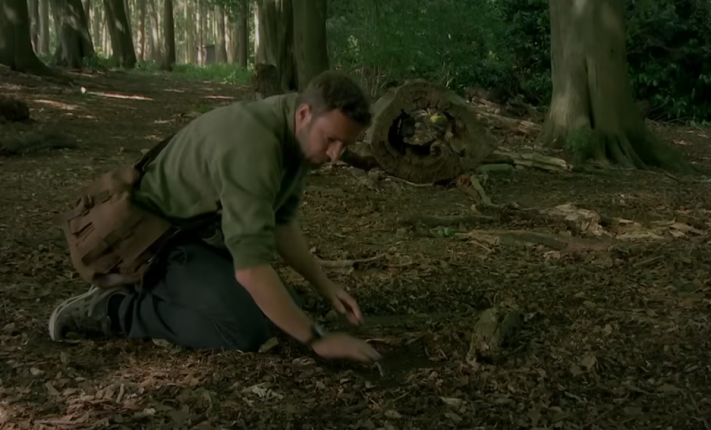 A man kneeling in a forest, examining the ground.