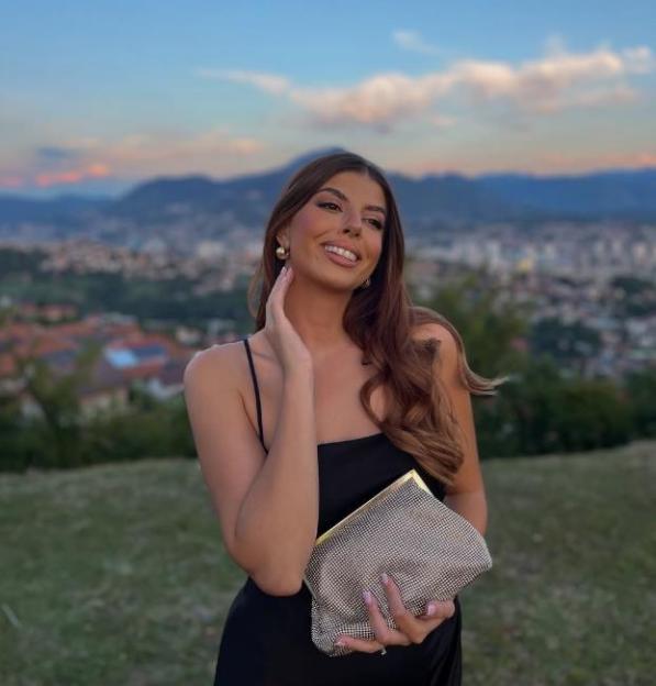 Adna, a young woman, in a black dress, smiling with a city skyline behind her.
