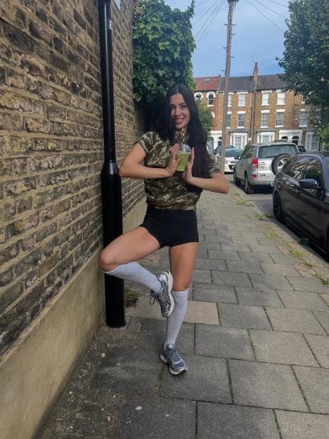 Woman holding a green smoothie, standing on one leg against a brick wall.