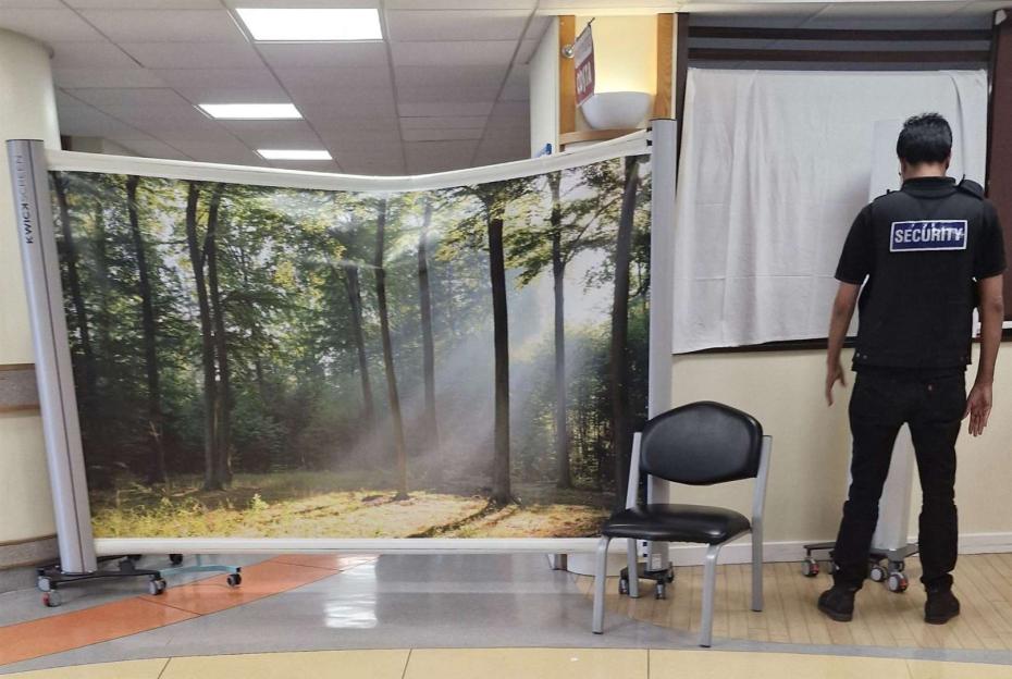 A security guard stands by a screen depicting a forest, blocking off the Costa café at William Harvey Hospital, which has been converted into a makeshift ward.