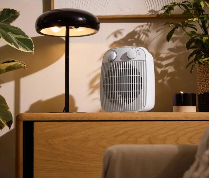 A white fan heater on a wooden table next to a black lamp.