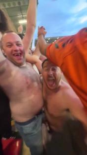 Two shirtless men celebrating enthusiastically amongst a crowd.