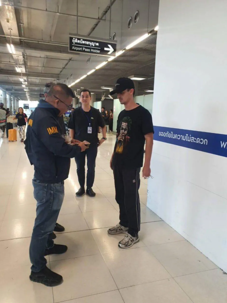 Three men standing in an airport terminal, one in a uniform with "IMM" on it, another in a black shirt, and a third in a black t-shirt and baseball cap.