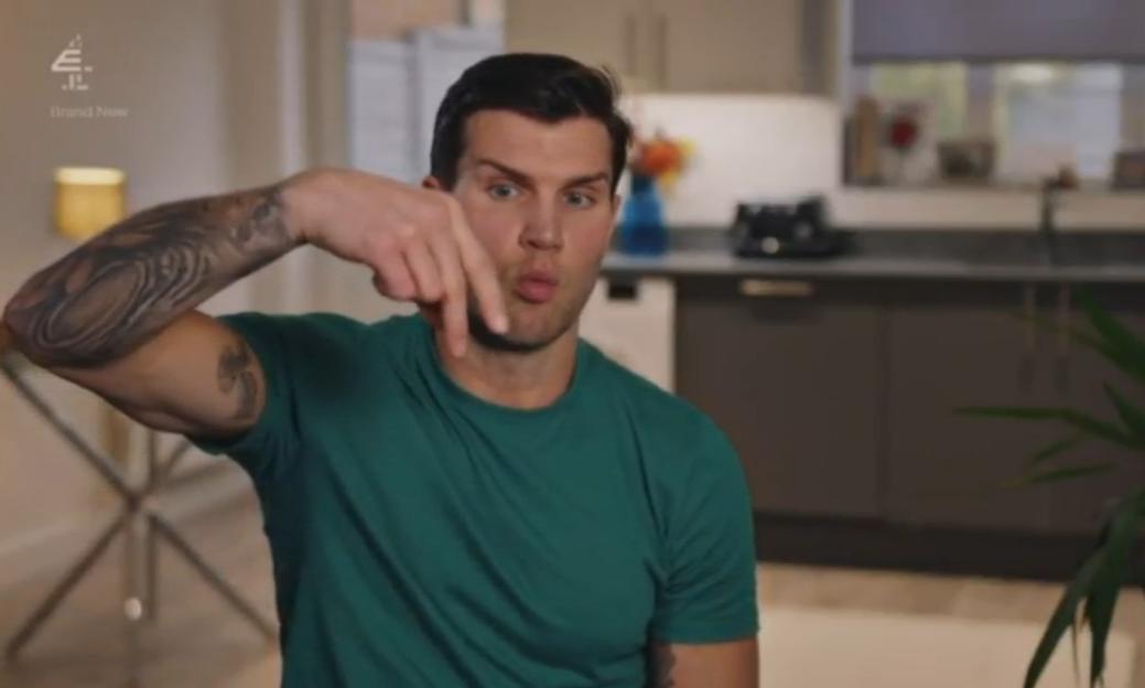 A man with tattooed arms in a teal t-shirt points downwards with both index fingers while making a face with pursed lips.