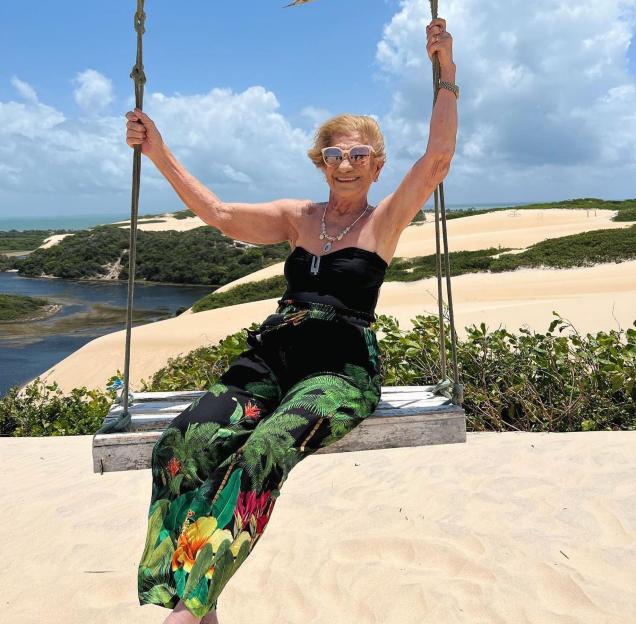 An 80-year-old woman sits on a swing overlooking a beach.