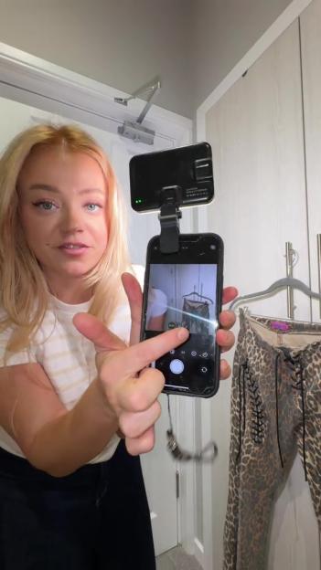 A blonde woman filming a video of an item of clothing hanging on a clothes rack with a smartphone with an attached light.