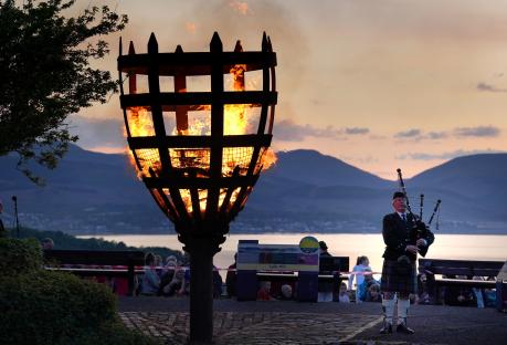 A large bonfire burning in a metal cage, with a bagpiper in a kilt playing in the foreground.