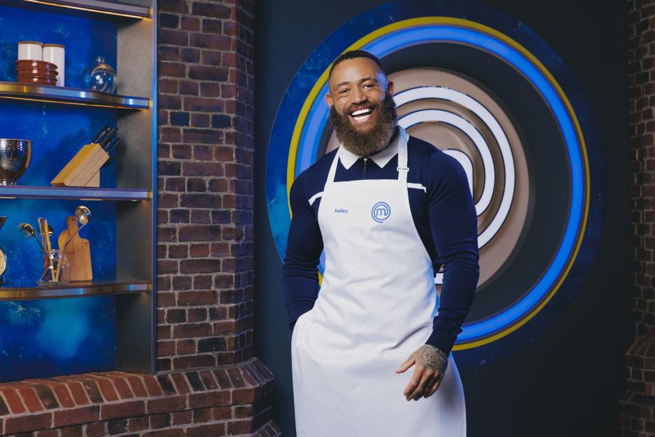 Ashley, a MasterChef contestant, poses in a blue and brick kitchen setting.