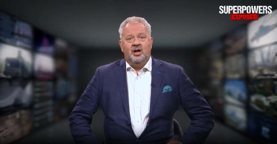 A white-haired, bearded man in a blue suit with a pocket square speaking on a show titled "Superpowers Exposed."