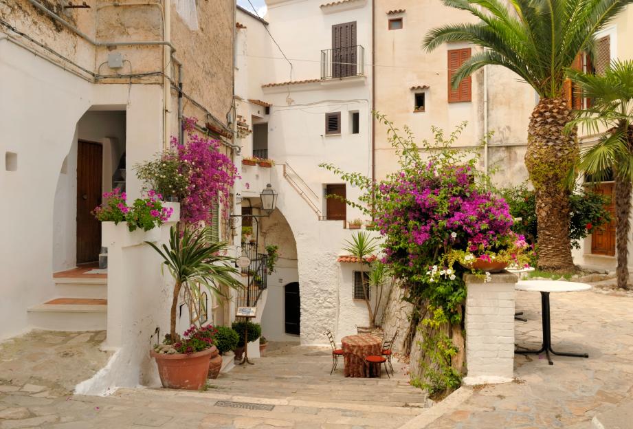 Street view of Sperlonga, Italy, with white buildings, flowering plants, and outdoor seating.