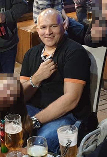 Geraint Berry, a bald man in a black polo shirt, black jeans, a large silver necklace, and a silver bracelet, smiles at the camera.